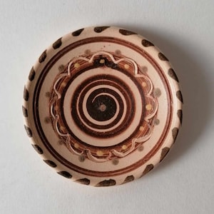 May include: A small, round, ceramic plate with a brown and white swirl design. The plate has a brown rim with a white border and a brown swirl in the center. The swirl is surrounded by a white border with a brown design.