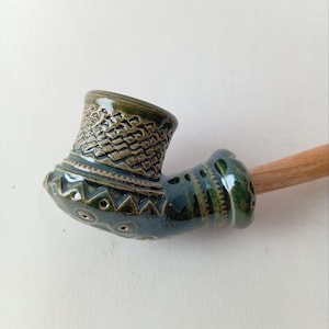 Puede incluir: Una pipa de cerámica hecha a mano con un esmalte verde oscuro. El cuenco presenta patrones texturizados y detalles decorativos. El tallo es de madera. Un accesorio para fumar único.