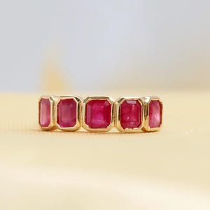 May include: A gold band ring featuring five rectangular, step-cut red rubies set in a row. Each ruby is encased in a smooth gold bezel setting, creating a continuous line of vibrant red gemstones across the top of the polished yellow gold band.