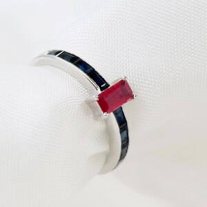 May include: A silver-toned ring featuring a central rectangular red gemstone set horizontally. The band is accented with a series of small, rectangular dark blue gemstones embedded along the shank, creating a contrast with the vibrant red center stone.