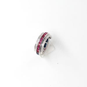 May include: A silver ring with three bands. The top band features small, clear stones, the middle band has red stones, and the bottom band has blue stones. The ring is displayed on a clear stand against a white background.