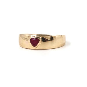 May include: A gold ring with a heart-shaped, deep red gemstone. The ring has a smooth, polished surface and a wide band. The heart-shaped stone is set into the band, creating a simple yet elegant design. The ring is likely made of gold.