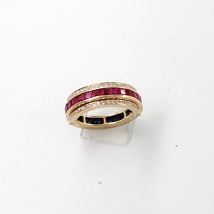 May include: A gold ring with three rows of gemstones. The top row features square-cut rubies, the middle row has small diamonds, and the bottom row has square-cut sapphires. The ring is set against a white background.