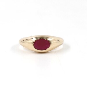 Può includere: Un anello d'oro con una fascia liscia e arrotondata e una gemma rosso scuro di forma ovale al centro. Il design dell'anello è semplice ed elegante.