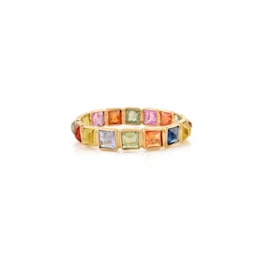 Puede incluir: Un anillo de oro con una serie de piedras preciosas de corte cuadrado en varios colores, como amarillo, naranja, rosa, verde y azul. Las piedras están muy juntas, creando una banda continua de color. El anillo se muestra sobre un fondo blanco.