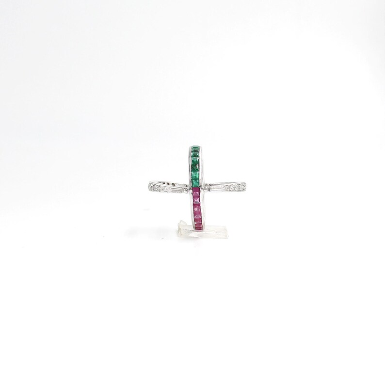 May include: A silver ring with a cross-shaped design. The ring features a row of green and red gemstones, with smaller white gemstones on the cross arms. The ring is set against a white background.