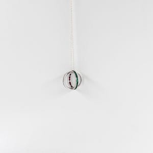 May include: A silver pendant necklace with a spherical design. The sphere is made of multiple rings, with alternating sections of green and purple gemstones. The necklace hangs from a delicate silver chain, creating an elegant and modern appearance.