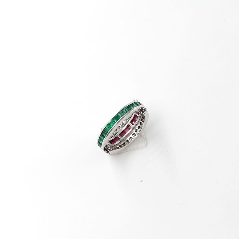 May include: A silver ring with a double band design. The top band is lined with green gemstones, while the inner band features red gemstones. The ring is set against a white background.