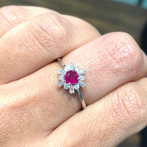 Può includere: Un anello in oro bianco con una pietra di rubino rotonda circondata da un alone di diamanti più piccoli.