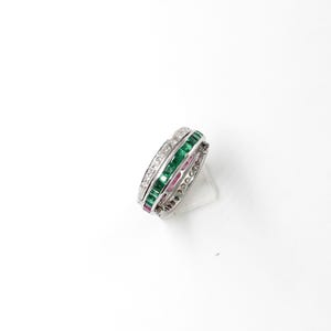 May include: A silver ring with three bands of gemstones. One band features clear stones, another emerald green stones, and the third band has pink stones. The ring is set against a white background.
