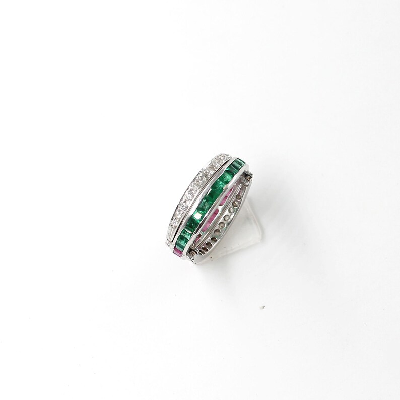 May include: A silver ring with three bands of gemstones. One band features clear stones, another emerald green stones, and the third band has pink stones. The ring is set against a white background.