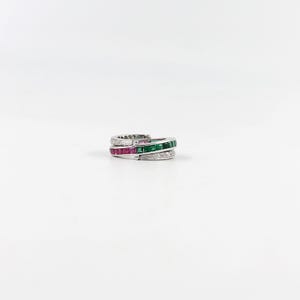 May include: A silver ring with three bands. The top band is encrusted with small white stones. The middle band features square-cut red stones, and the bottom band has square-cut green stones. The ring is set against a white background.