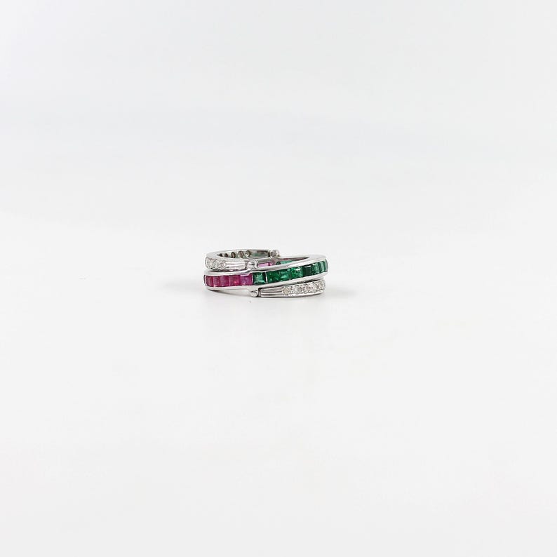 May include: A silver ring with three bands. The top band is encrusted with small white stones. The middle band features square-cut red stones, and the bottom band has square-cut green stones. The ring is set against a white background.