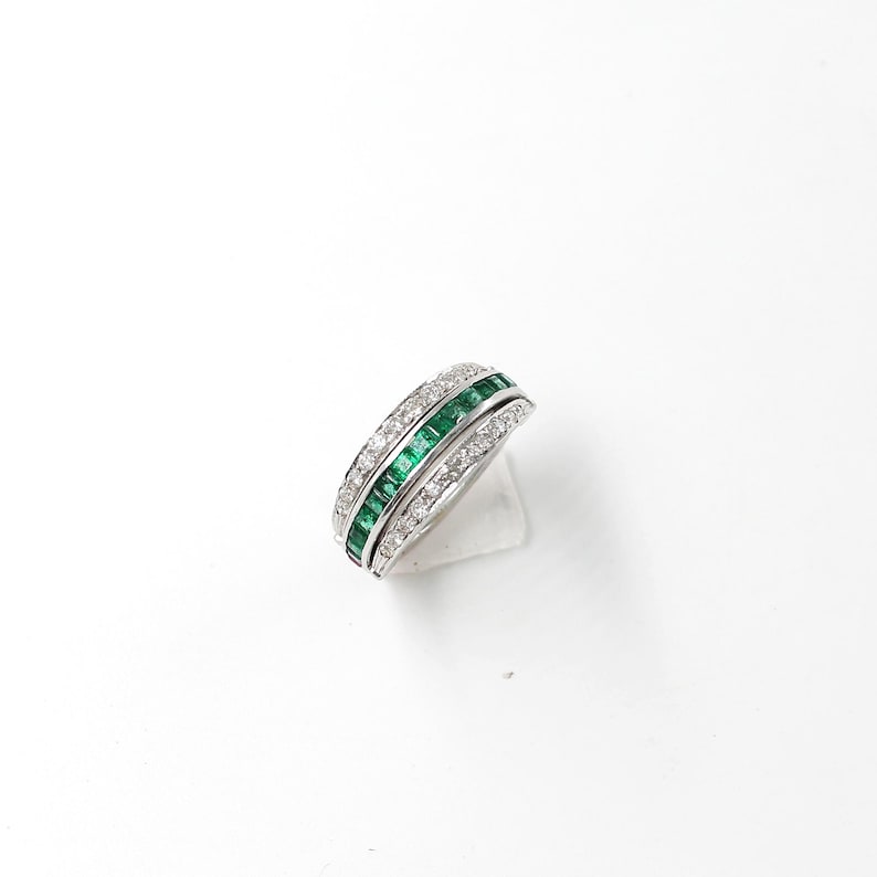 May include: A silver ring featuring two bands of emerald-cut green gemstones and a band of small, clear gemstones. The ring is displayed on a white surface, highlighting its elegant design and sparkling details.