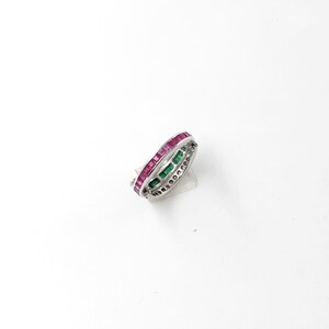 May include: A silver ring with three rows of gemstones. The top row features rectangular red stones, the middle row has green stones, and the bottom row is adorned with small, clear stones. The ring is set against a white background.