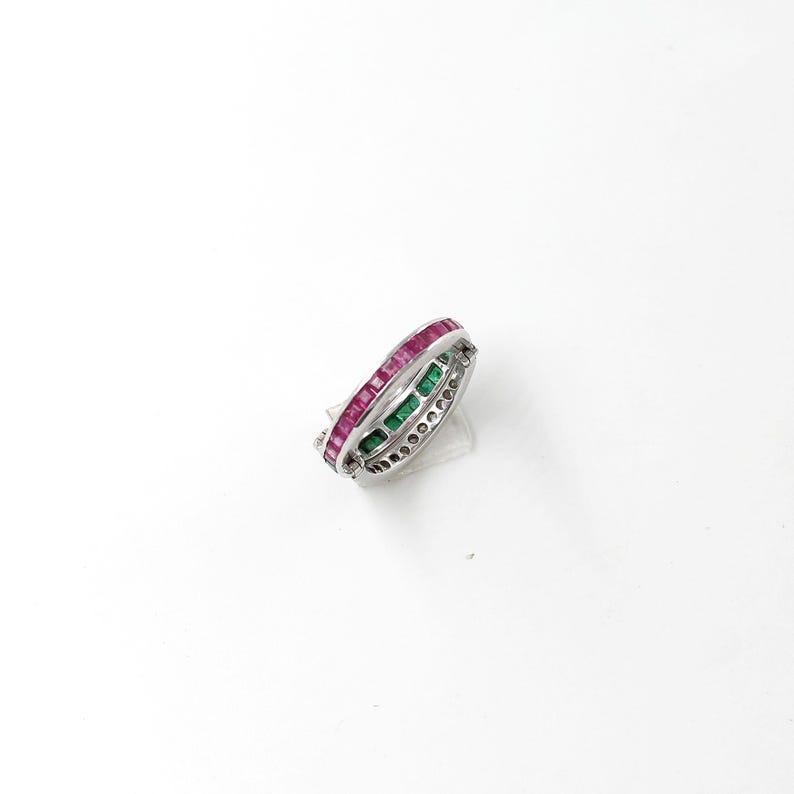 May include: A silver ring with three rows of gemstones. The top row features rectangular red stones, the middle row has green stones, and the bottom row is adorned with small, clear stones. The ring is set against a white background.