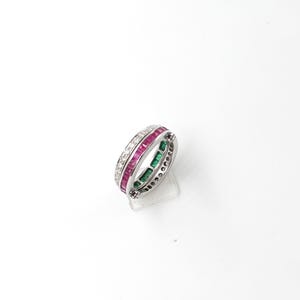 May include: A silver ring with three bands of gemstones. One band features small, square-cut rubies, another has emeralds, and the third is adorned with small diamonds. The ring is set against a white background.