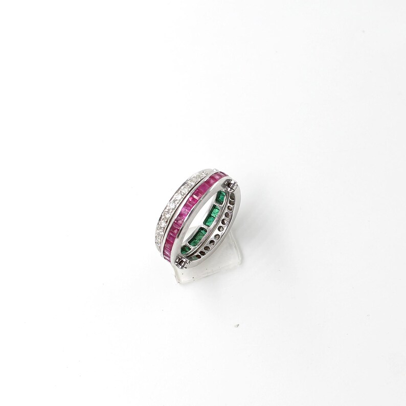May include: A silver ring with three bands of gemstones. One band features small, square-cut rubies, another has emeralds, and the third is adorned with small diamonds. The ring is set against a white background.
