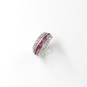 May include: A silver ring featuring three bands. The top and bottom bands are set with small, clear stones. The central band is filled with rectangular, deep red stones. The ring is shown on a white surface.