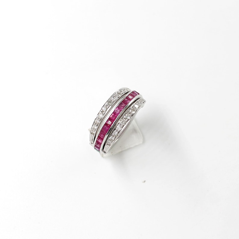 May include: A silver ring featuring three bands. The top and bottom bands are set with small, clear stones. The central band is filled with rectangular, deep red stones. The ring is shown on a white surface.
