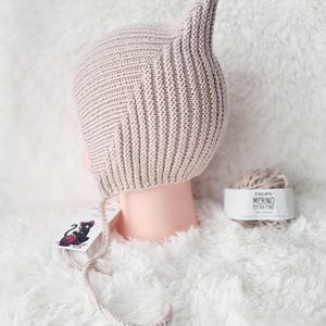Könnte beinhalten: Eine hellbeige gestrickte Mütze mit spitzer Spitze, auf einem Schaufensterpuppenkopf präsentiert. Die Mütze hat Ohrenklappen und Bindebänder. Ein Knäuel Wolle und ein Etikett mit einer schwarzen Katzenillustration sind ebenfalls sichtbar.