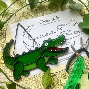 May include: A green stained glass crocodile suncatcher with a red mouth and a black chain. The design is next to a printed pattern and tools, suggesting a DIY craft project. The text "Crocodile" is visible.
