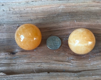 May include: Two smooth, yellow-orange, polished, crystal spheres on a wooden surface. The spheres are about the size of a US quarter.
