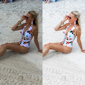 May include: A woman wearing a white and floral print swimsuit with a plunging neckline. She is sitting on a sandy beach with her sunglasses on.