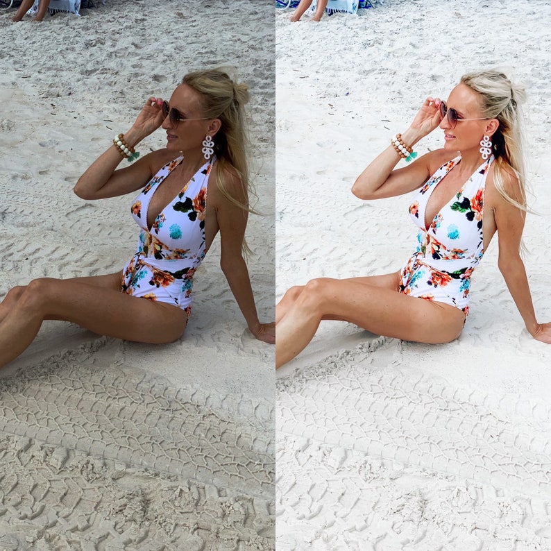 May include: A woman wearing a white and floral print swimsuit with a plunging neckline. She is sitting on a sandy beach with her sunglasses on.