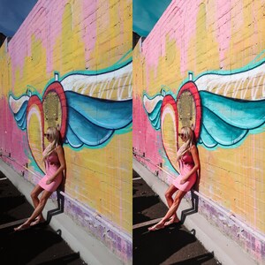 May include: A woman in a pink dress stands in front of a colorful wall with a heart and wings painted on it. The wall is painted in shades of pink, yellow, and blue.