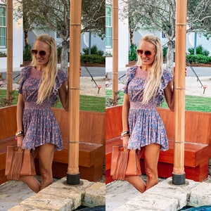 May include: A woman wearing a floral print dress and sunglasses stands in front of a wooden bench. She is holding a brown fringed bag.