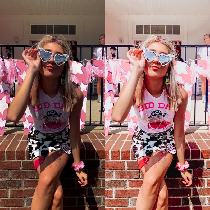May include: A woman wearing a white tank top with a graphic print of a cowboy hat and the text "Bid Day" on it. She is also wearing a black and white cow print skirt, heart-shaped sunglasses, and a pink headband. She is standing in front of a brick wall with pink and white decorations.
