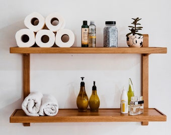 May include: Two wooden shelves against a white wall. The top shelf has five rolls of toilet paper, three bottles of liquid soap, a jar of bath salts, and a small potted plant. The bottom shelf has two bottles of liquid soap, a roll of towels, a bottle of lotion, and a bottle of perfume.