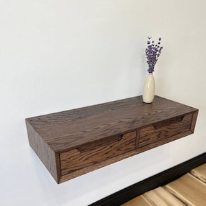 Puede incluir: Una estantería flotante de madera marrón oscuro con dos cajones. La estantería está montada en una pared blanca y tiene un jarrón con flores de lavanda secas en la parte superior.
