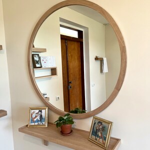 May include: A round wooden framed mirror with a shelf underneath. The shelf has two framed pictures and a small potted plant. The mirror reflects a hallway with a wooden door.