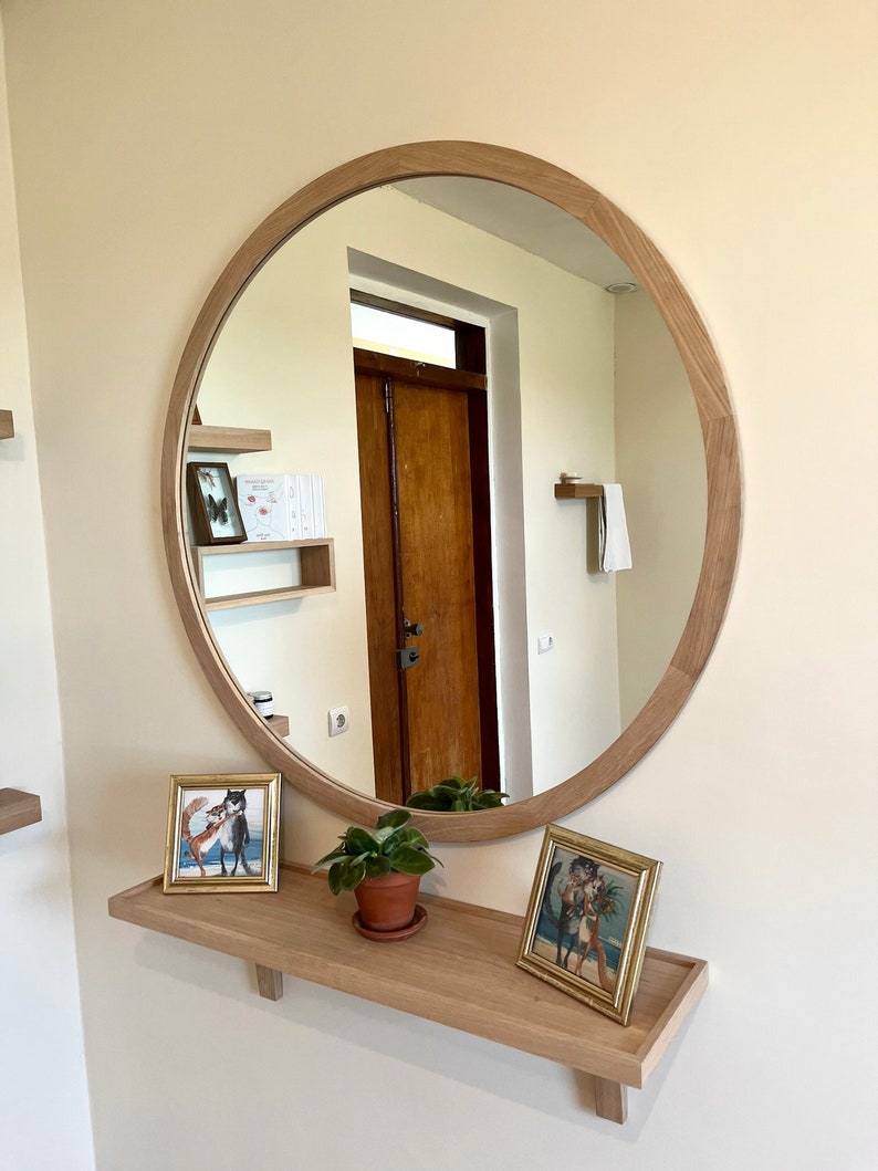 May include: A round wooden framed mirror with a shelf underneath. The shelf has two framed pictures and a small potted plant. The mirror reflects a hallway with a wooden door.