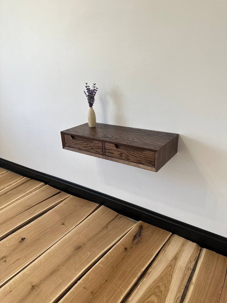May include: A dark wood floating shelf with two drawers. The shelf is mounted on a white wall and has a small vase with purple flowers on top.