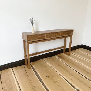 May include: A wooden console table with three drawers and a simple design. The table is made of light brown wood and has a natural finish. It is standing on a wooden floor.
