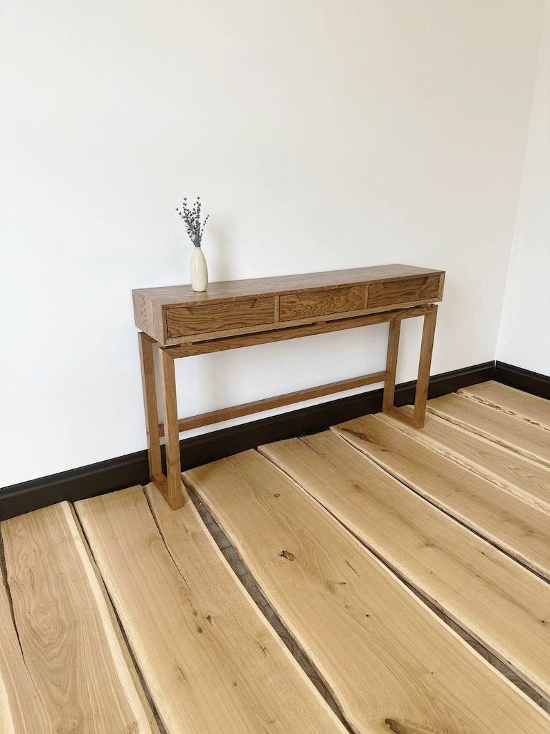 May include: A wooden console table with three drawers and a simple design. The table is made of light brown wood and has a natural finish. It is standing on a wooden floor.