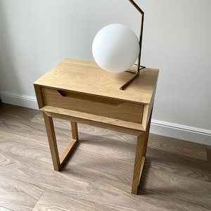 May include: A light wood nightstand with a single drawer and a gold metal table lamp with a white globe. The nightstand is on a light wood floor.