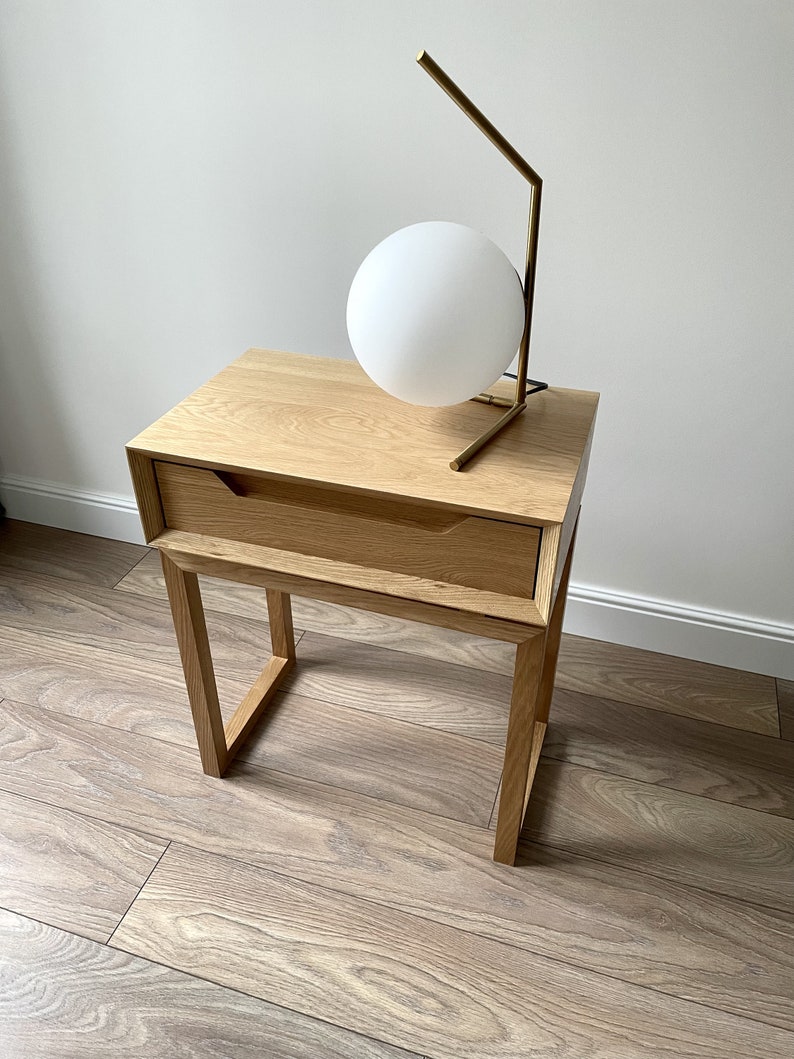 May include: A light wood nightstand with a single drawer and a gold metal table lamp with a white globe. The nightstand is on a light wood floor.