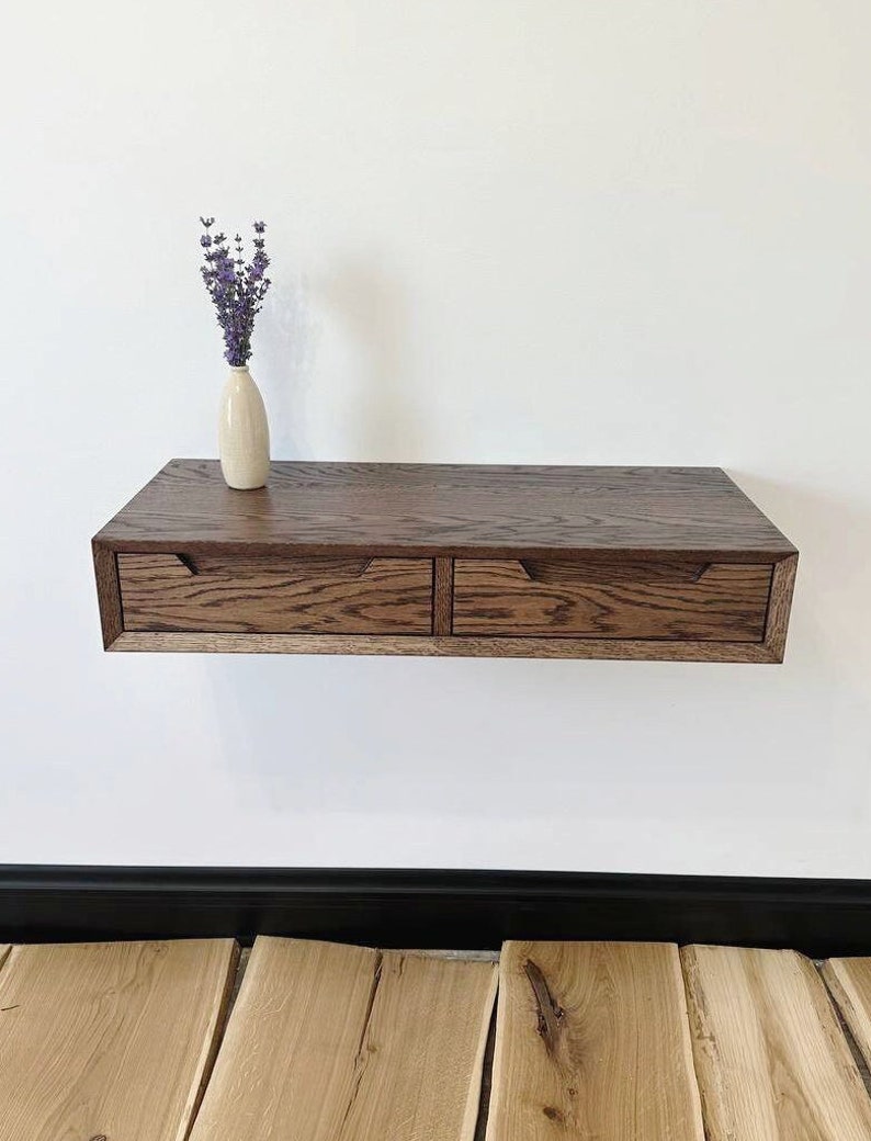 May include: A floating wall shelf with two drawers made of dark wood. The shelf is mounted on a white wall and has a vase with purple flowers on top.