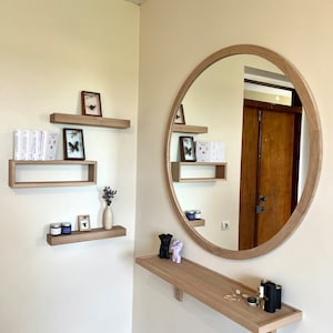 May include: A round wooden mirror with a light wood frame hangs on a wall with a shelf underneath. The shelf is decorated with two small black figurines, a small bottle, and a few pieces of jewelry. There are three shelves above the mirror, each with a small framed picture or a collection of books.