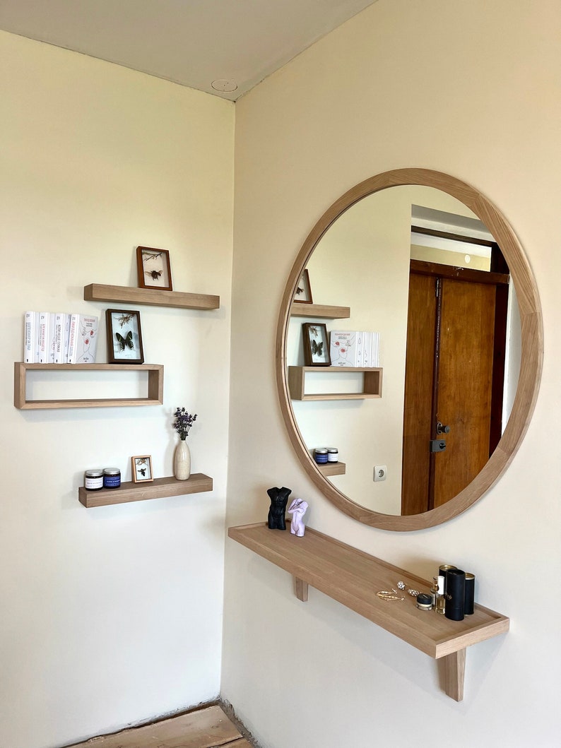 May include: A round wooden mirror with a light wood frame hangs on a wall with a shelf underneath. The shelf is decorated with two small black figurines, a small bottle, and a few pieces of jewelry. There are three shelves above the mirror, each with a small framed picture or a collection of books.