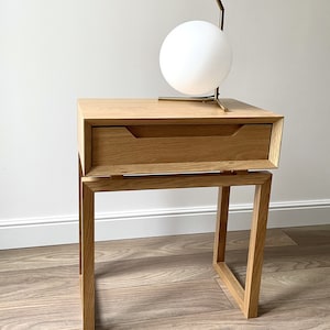 May include: A wooden nightstand with a single drawer and a metal lamp with a white globe on top. The nightstand has a simple, modern design and is made of light wood.