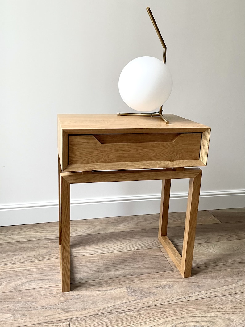 May include: A wooden nightstand with a single drawer and a metal lamp with a white globe on top. The nightstand has a simple, modern design and is made of light wood.