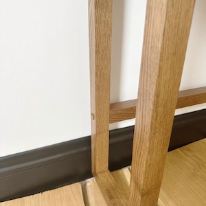 May include: Close-up of the bottom of a wooden table leg. The leg is made of light brown wood and has a simple, rectangular design. The table leg is resting on a wooden floor.
