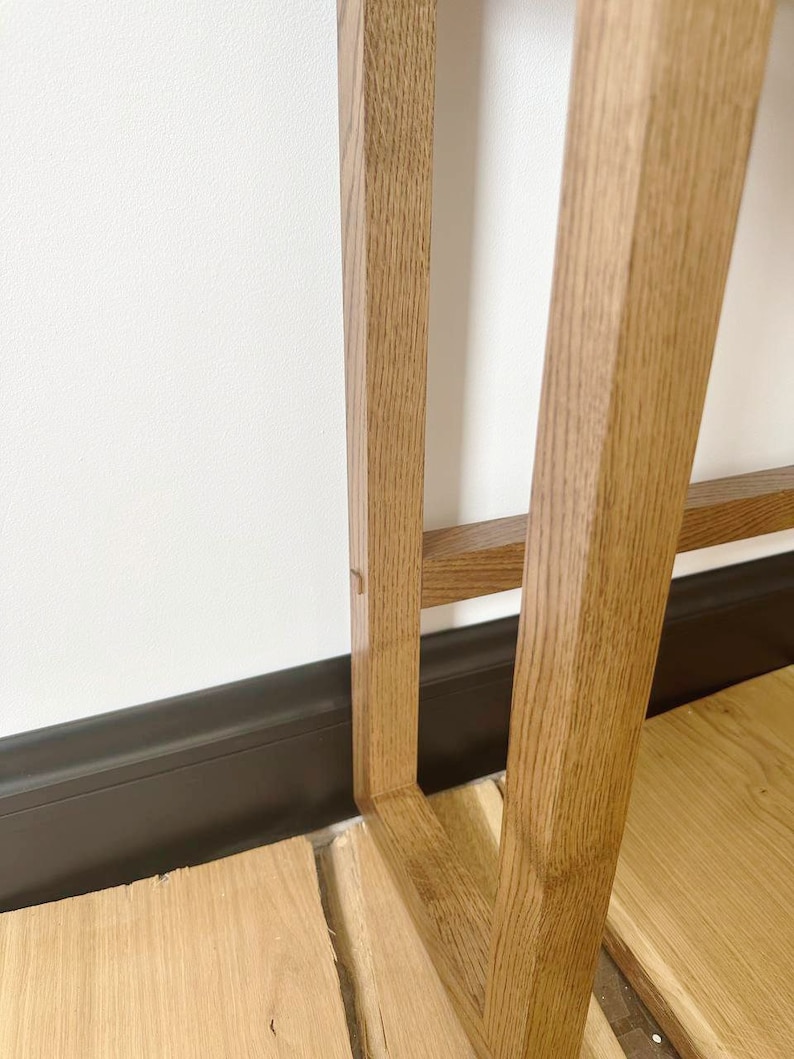 May include: Close-up of the bottom of a wooden table leg. The leg is made of light brown wood and has a simple, rectangular design. The table leg is resting on a wooden floor.