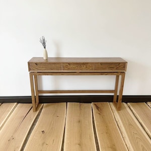 May include: A wooden console table with three drawers and a simple, modern design. The table is made of light brown wood and has a natural finish. It is sitting on a wooden floor.