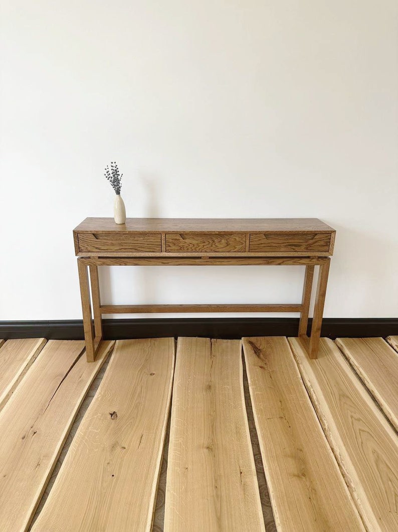May include: A wooden console table with three drawers and a simple, modern design. The table is made of light brown wood and has a natural finish. It is sitting on a wooden floor.
